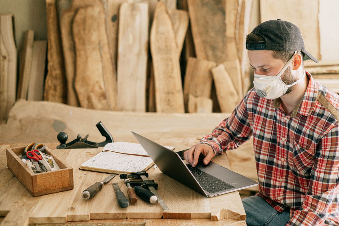 A craftsman wearing a dust mask checks a laptop screen in his woodworking shop, representing a small maker business that benefits from automated monitoring.